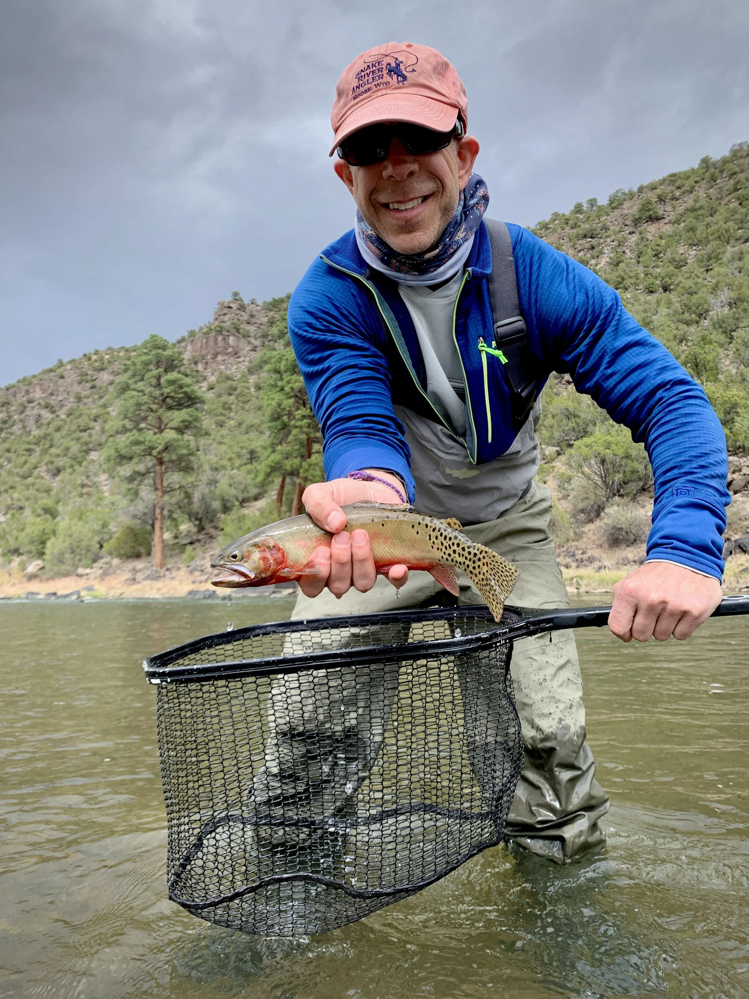 A man in outdoor fishing gear holding a caught fish and a fishing net in a river with trees and mountains in the background.