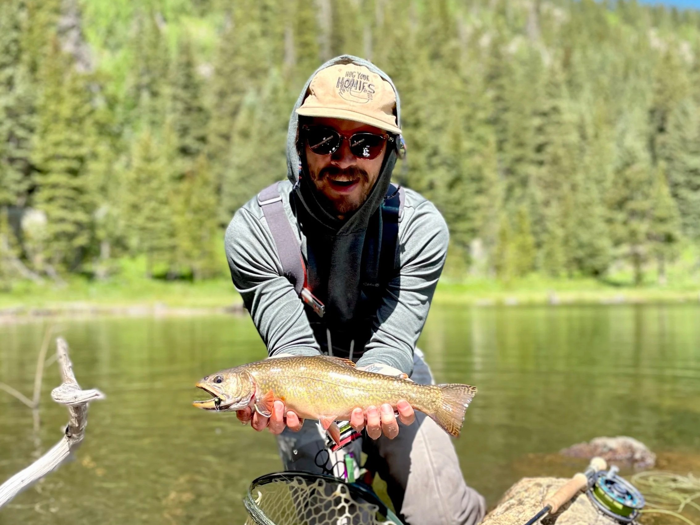 Man wearing sunglasses, a hat with the words 'HOG YOUR HOMIES', and a hoodie, holding a fish by the water in a forested area, smiling at the camera.