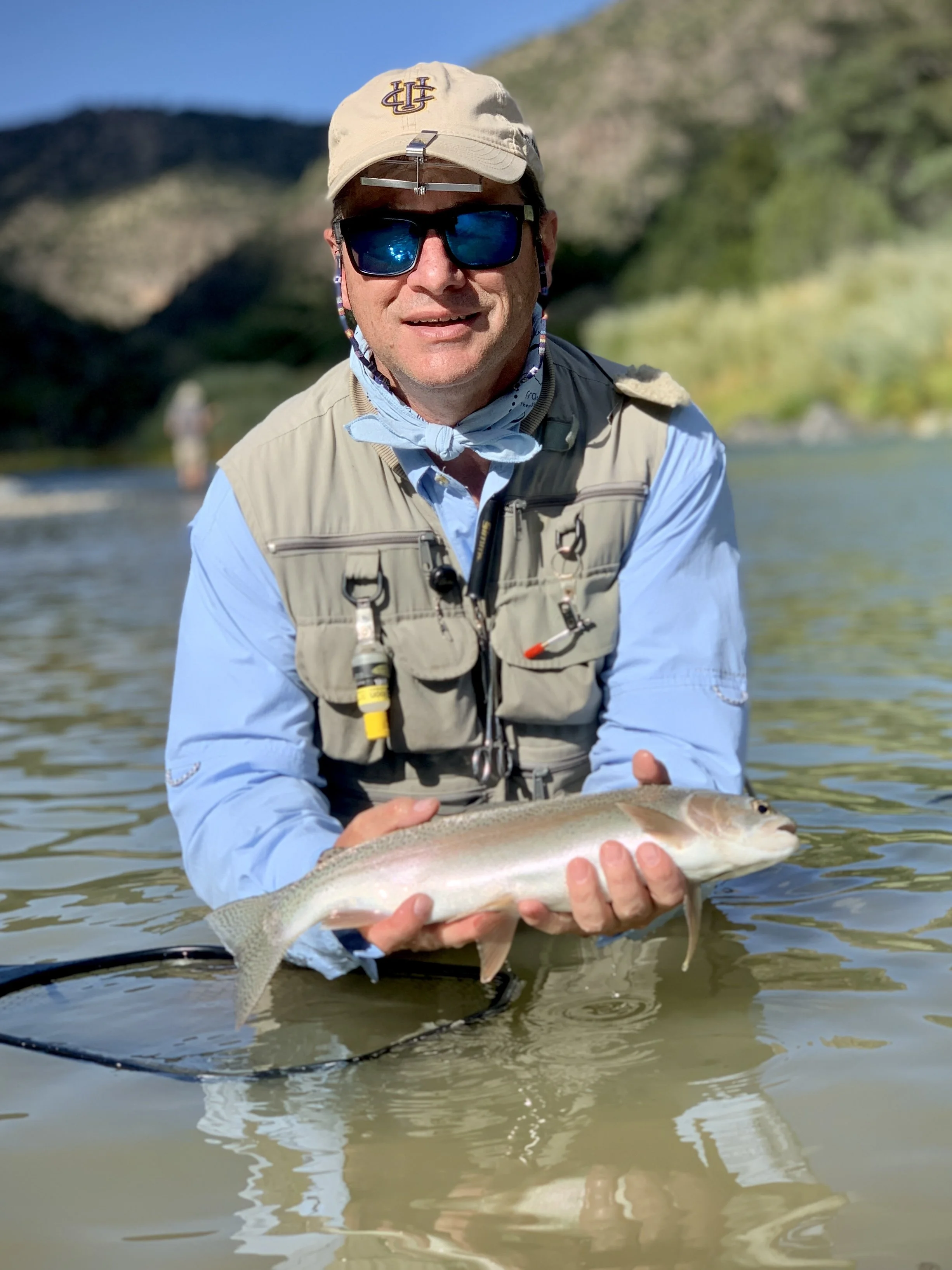 Man in sunglasses, hat, and fishing gear holding a fish in a river during daytime, with mountains and trees in the background.