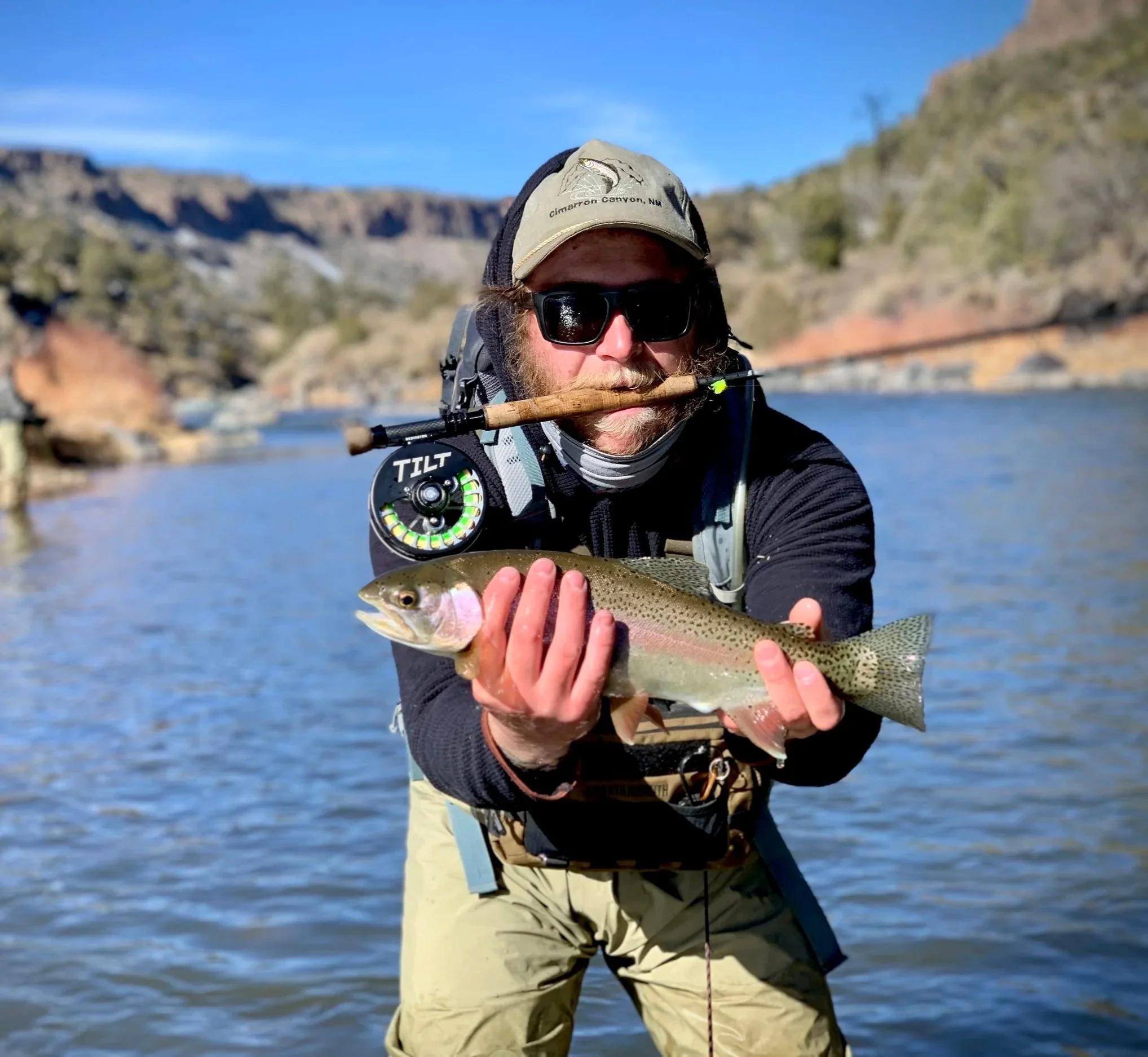 Man fishing with a fly fishing rod, holding a rainbow trout he caught, in a river with a mountainous background.