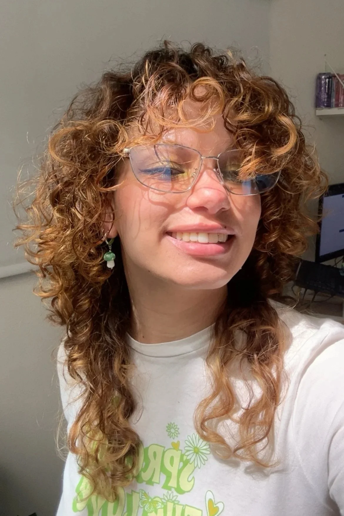 Une femme aux cheveux bouclés et roux, portant des lunettes et un t-shirt blanc avec des motifs verts, sourit dans une pièce avec un ordinateur en arrière-plan.