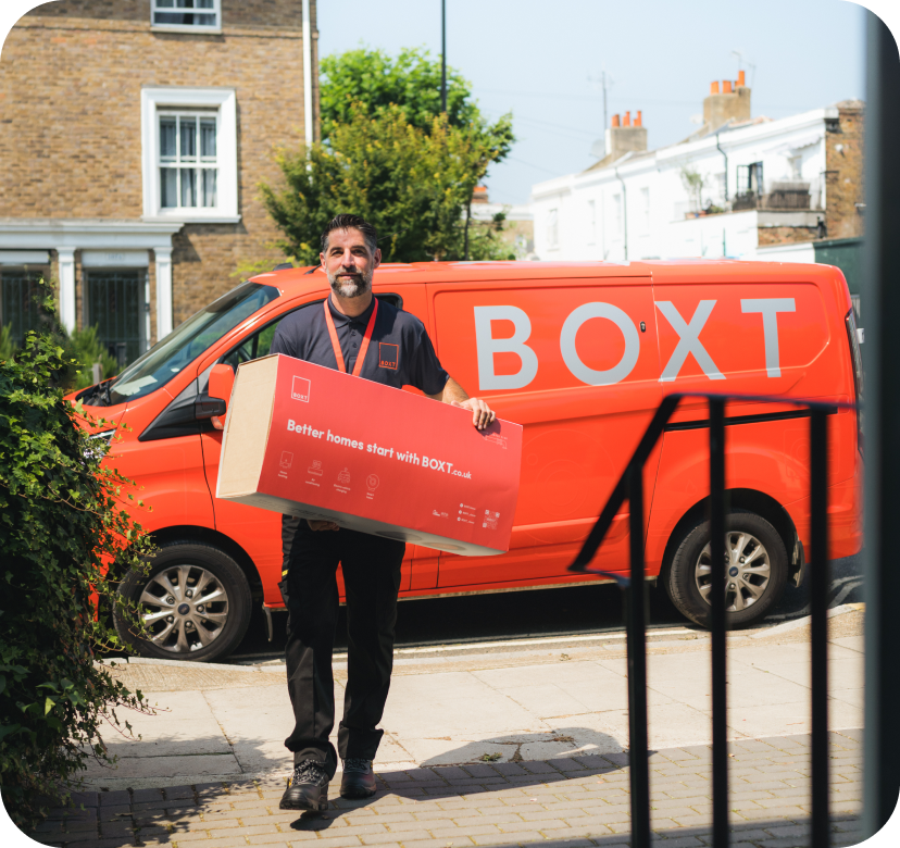 A BOXT engineer holding a large box while standing outside in front of a red BOXT delivery van with the logo and slogan. The scene is set in a residential neighbourhood.
