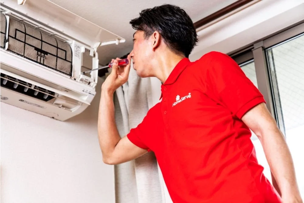 A HomeServe engineer in a red uniform is inspecting an air conditioning unit mounted on the wall inside a room.