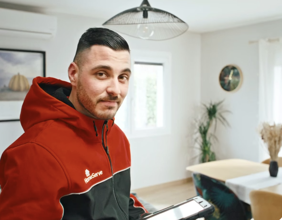 HomeServe engineer in a red jacket sitting in a bright dining room, holding a tablet, with a window, plants, and wall decor in the background.