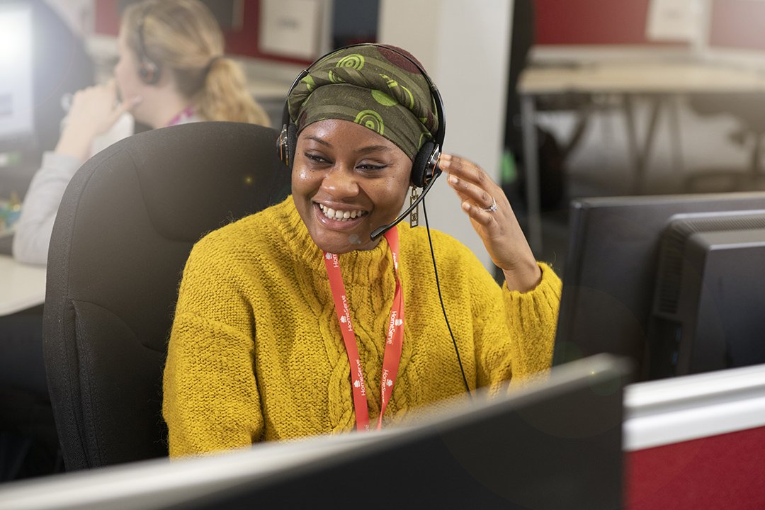 A HomeServe customer service agent wearing a green headscarf and a yellow sweater, smiling while working at her desk with a headset on.