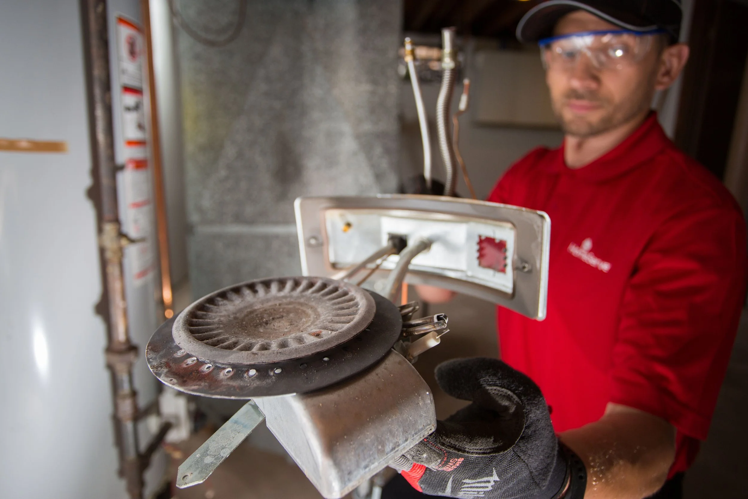 A technician in safety glasses, black gloves, and a red shirt holding a removed electric stove coil in an indoor setting with a water heater and pipes in the background.