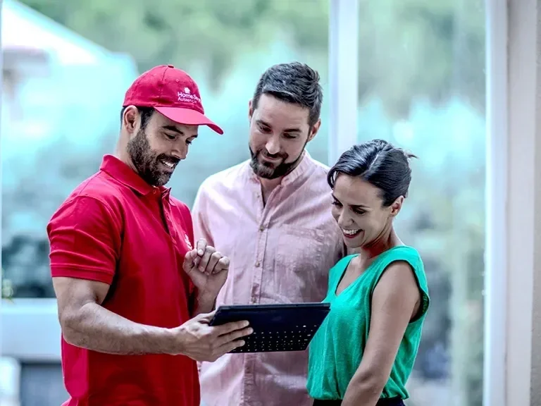 A HomeServe engineer wearing a red cap and polo shirt showing something on a tablet to a couple, all smiling and looking at the screen in a brightly lit indoor space.