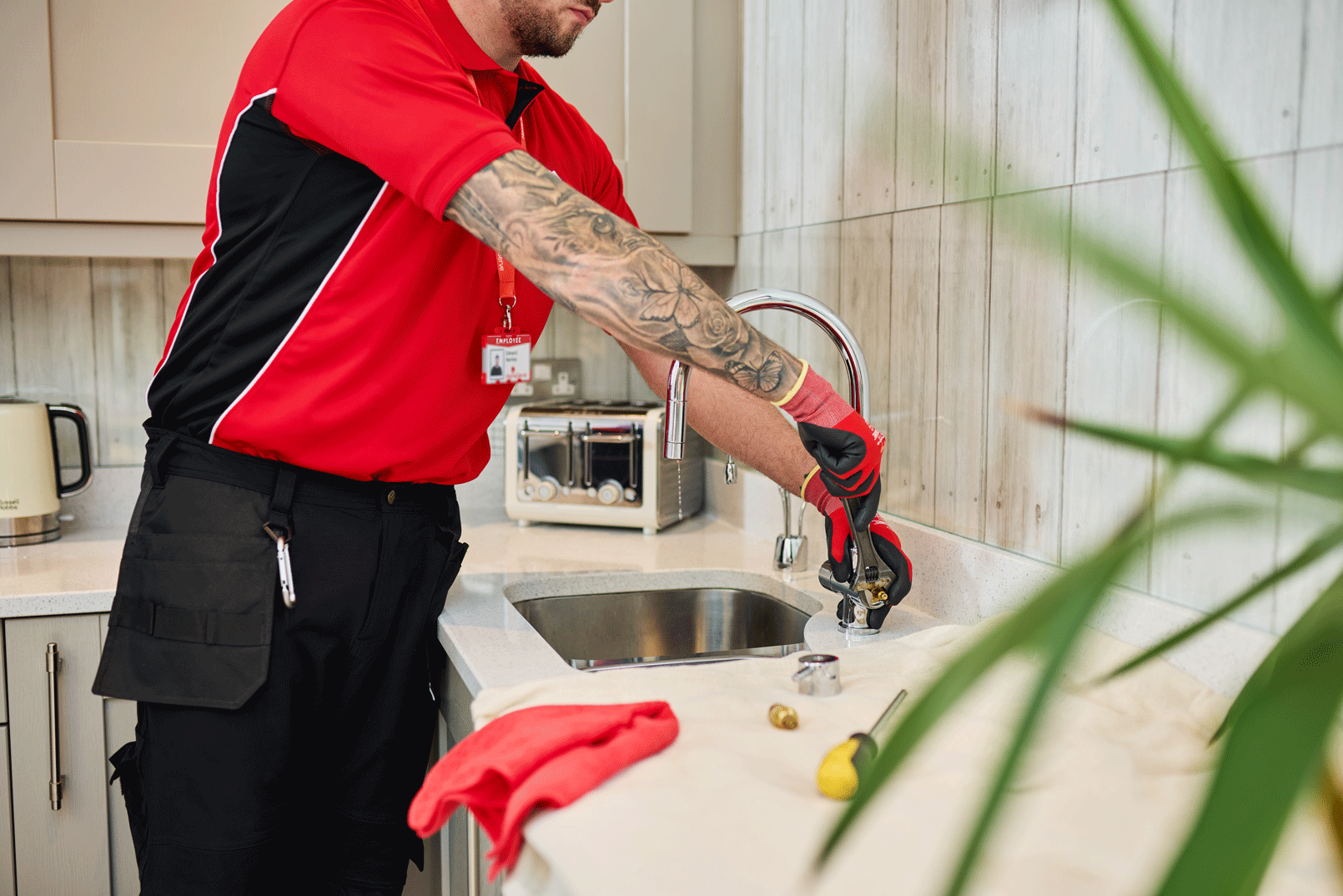 A HomeServe engineer is working on fixing a kitchen sink faucet. The man has a tattooed arm and is wearing red gloves. The kitchen has a beige countertop with a towel and a screwdriver on it, and kitchen appliances are visible in the background.
