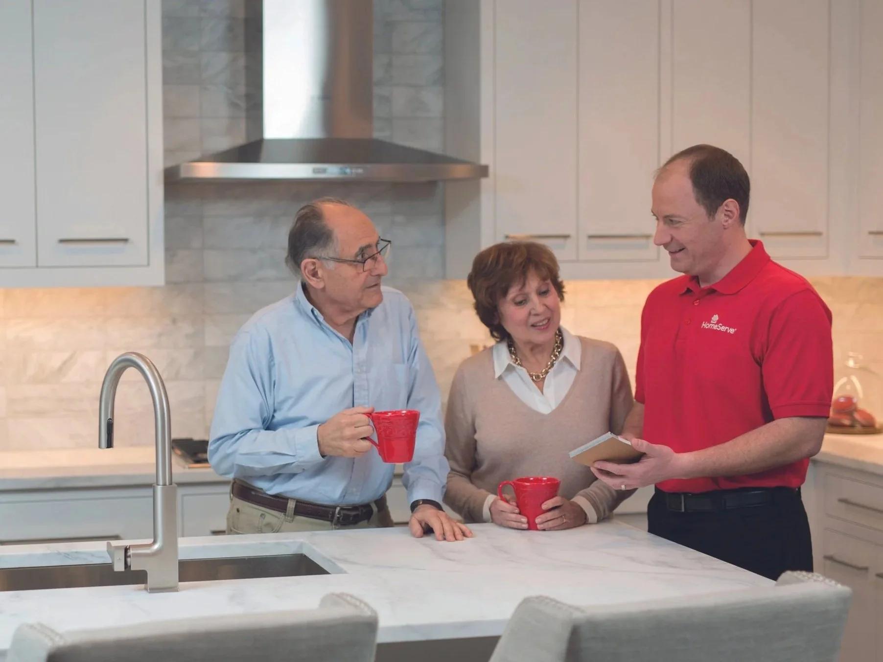 Three adults standing in a kitchen, engaged in conversation, holding red coffee mugs, with a HomeServe engineer showing a iPad to an older couple.