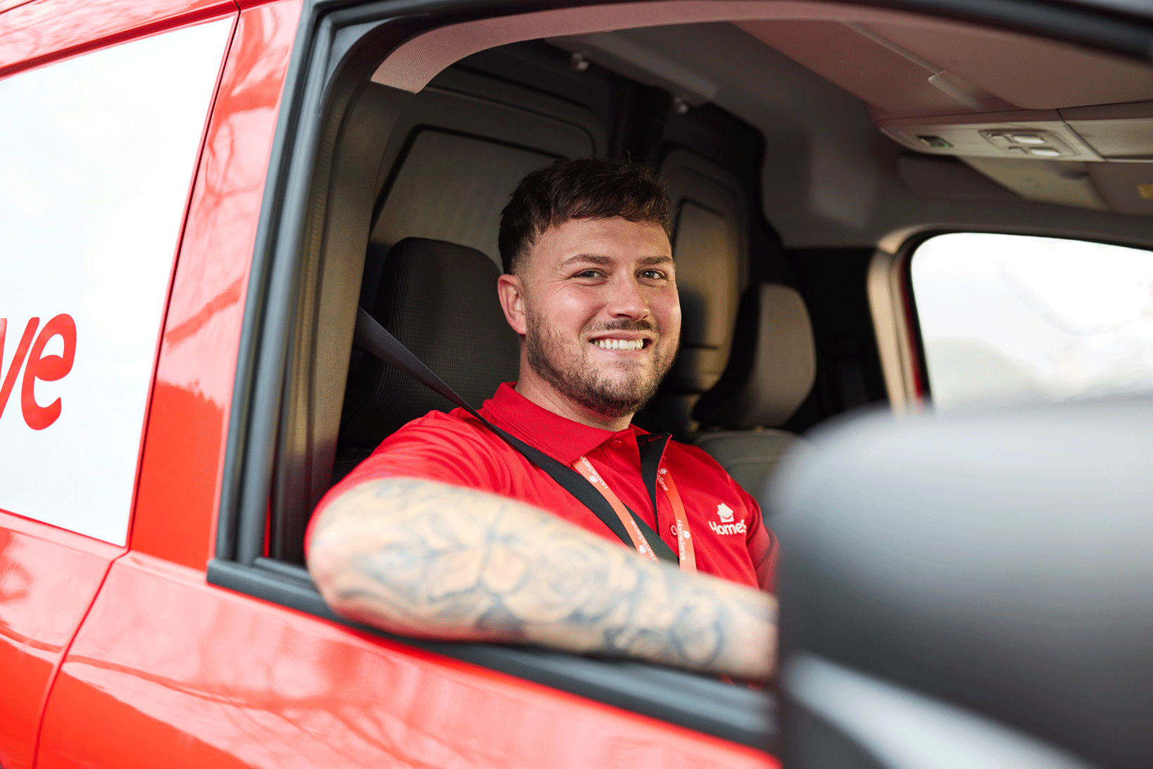 A smiling HomeServe engineer with curly hair wearing a red polo shirt with 'HomeServe' logo, holding a coffee mug, standing in a modern kitchen with dark cabinets and a white marble countertop.