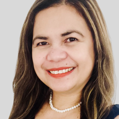 Smiling woman with light brown hair, wearing a pearl necklace and a dark top, against a plain light background.