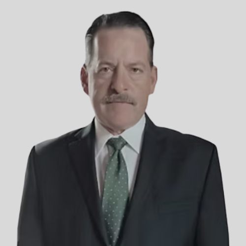 A middle-aged man in a suit with a green tie and mustache, standing against a plain white background.