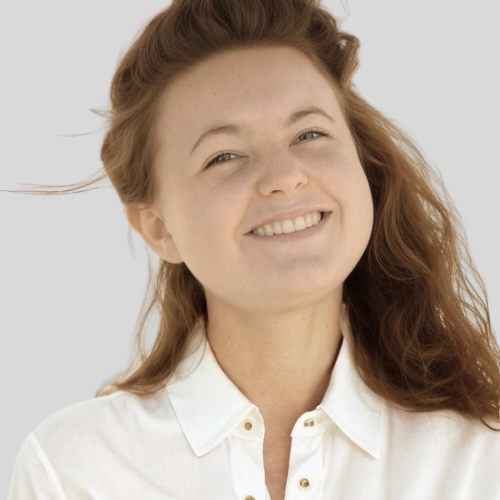 A young woman with long, wavy red hair smiling, wearing a white collared shirt, against a plain light background.