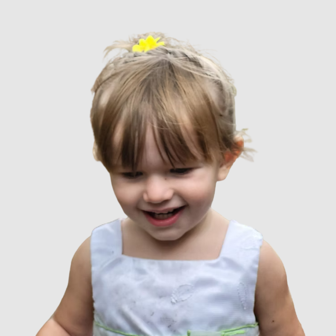 A young girl with short light brown hair, wearing a white dress with green accents and a yellow flower hair clip, smiling and looking down.
