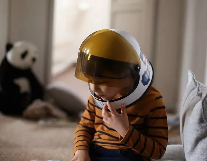 A young boy wearing a striped orange and black shirt trying on a space helmet in a living room with a panda plush toy in the background.