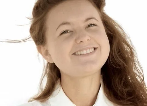 A woman with light skin and red hair smiling at the camera, wearing a white shirt against a white background.