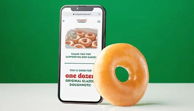 A smartphone displaying a website for Krispy Kreme, with a glazed donut on the right side of the phone against a green background.