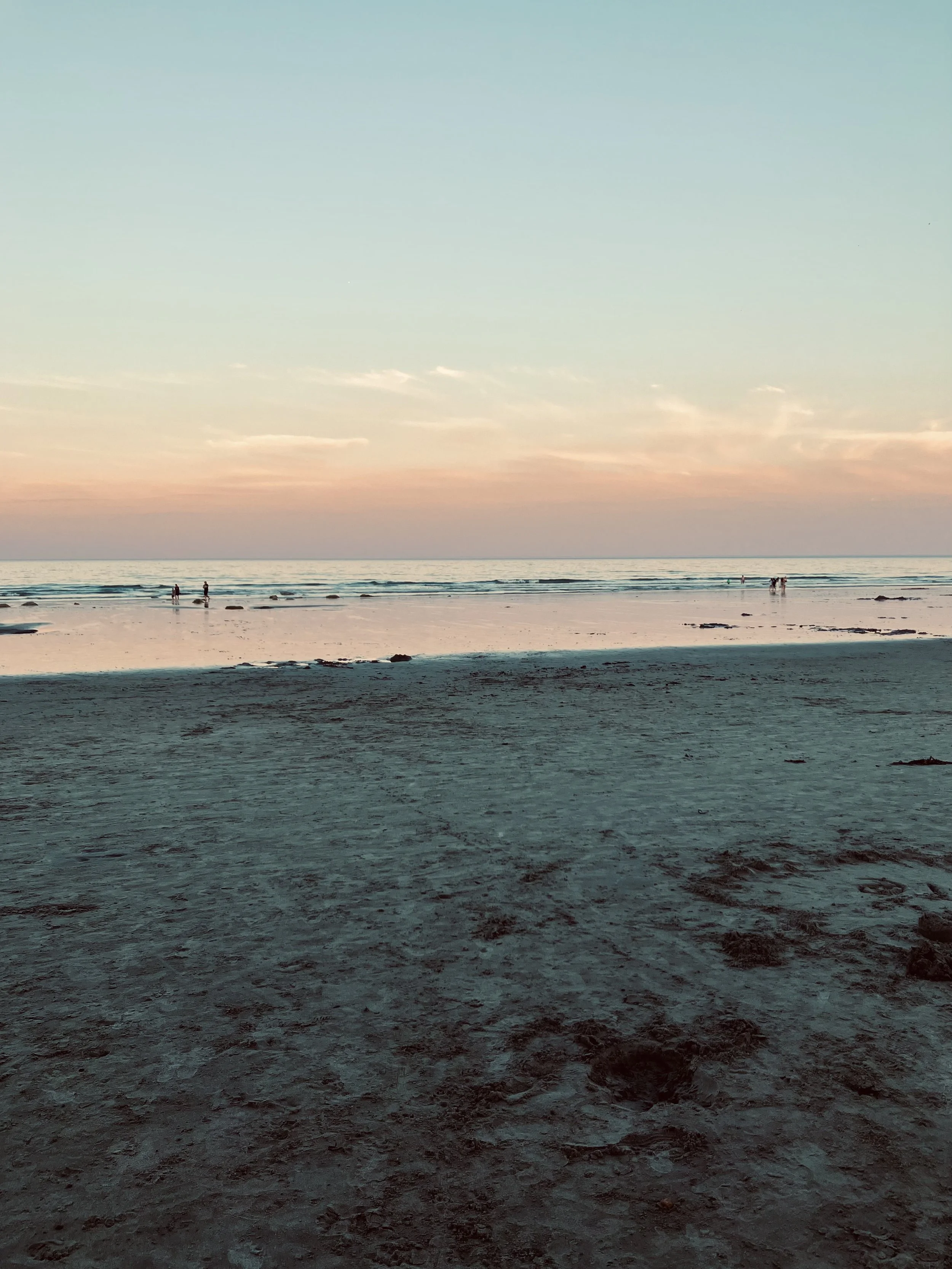 Beach scene during sunset or sunrise with a clear sky, calm ocean waves, and a few people walking along the shore.
