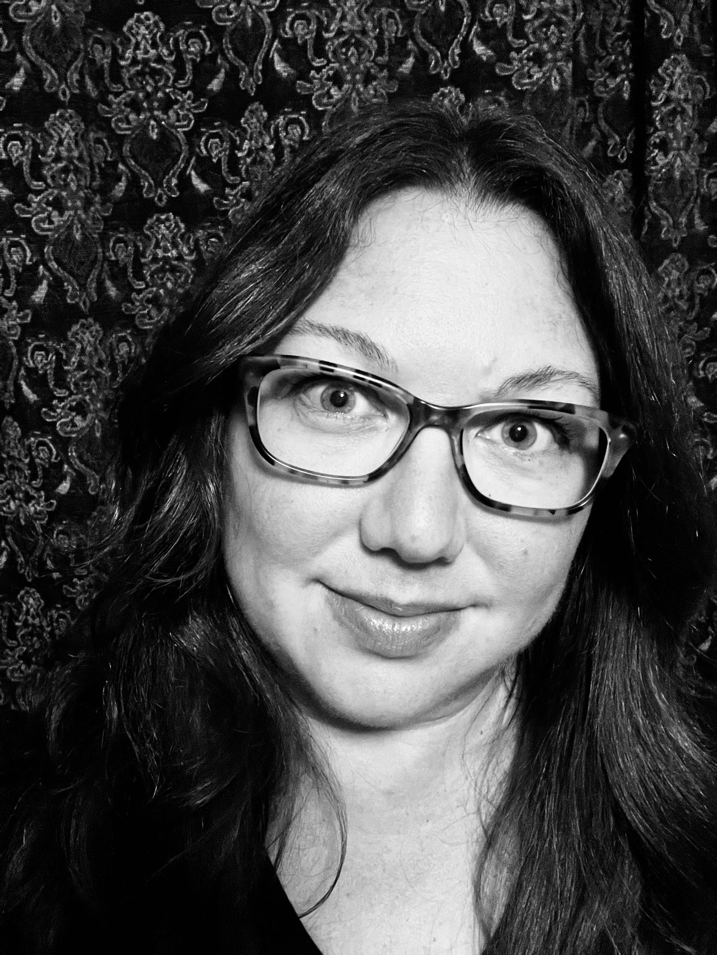 Black and white portrait of a woman with long wavy hair, wearing glasses, smiling, with a patterned background.