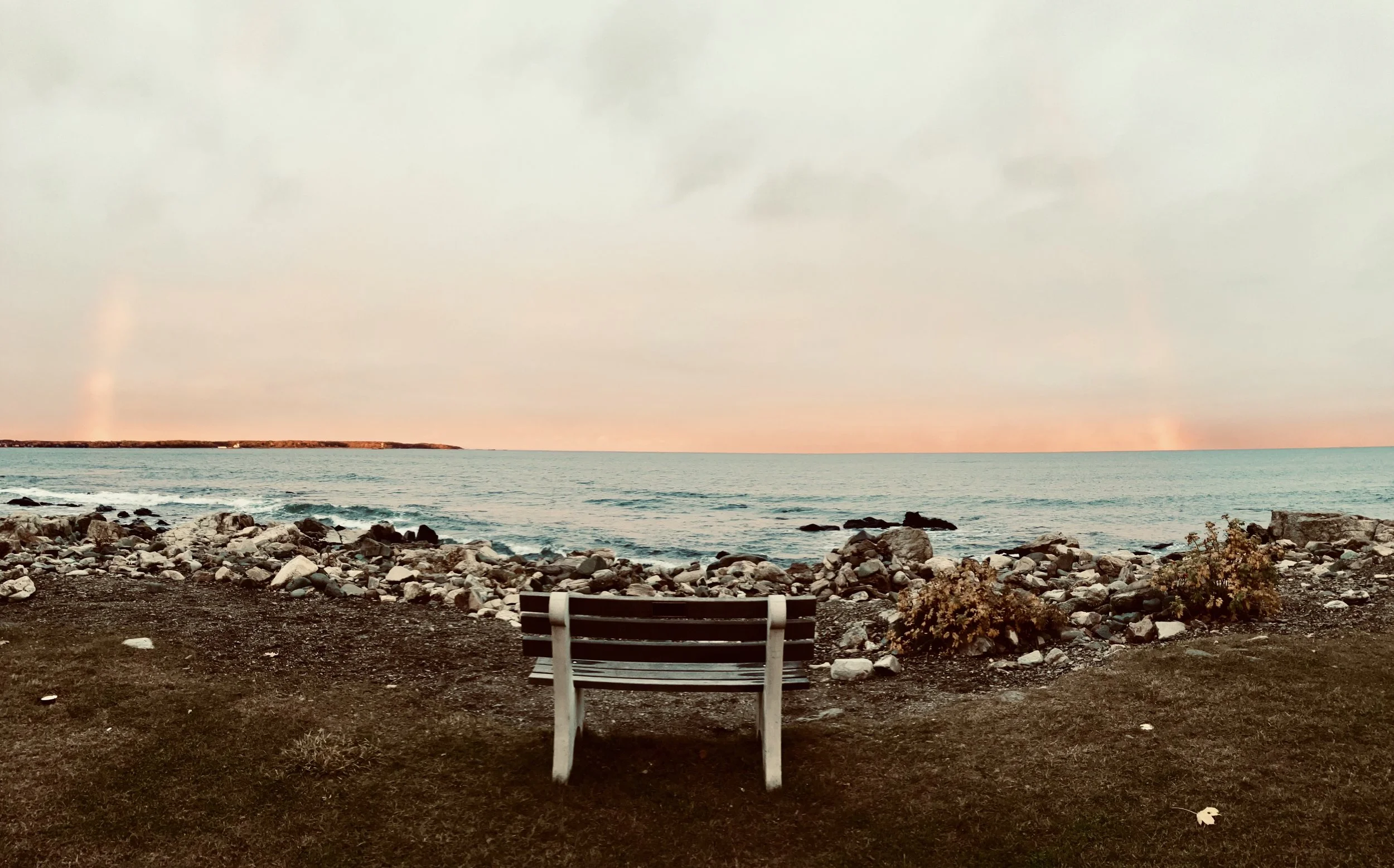 A wooden bench facing the ocean on a rocky shoreline at sunset with a cloudy sky.