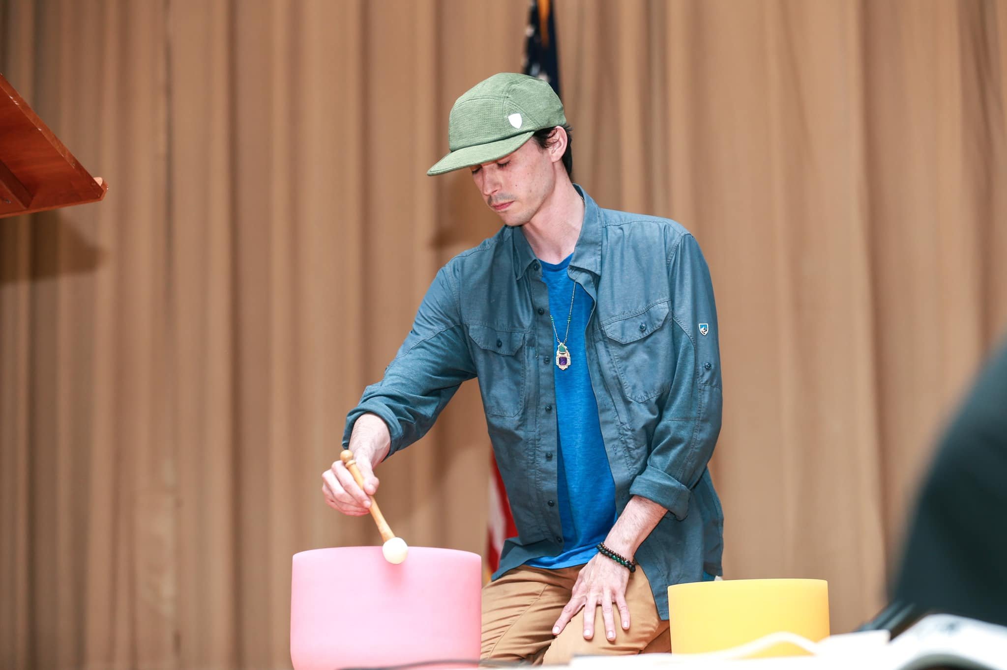 A man wearing a green cap, blue shirt, and tan pants playing a pink steel drum with a mallet inside a room with wood-paneled walls and an American flag in the background.