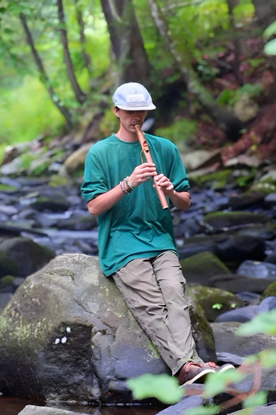A young man wearing a gray cap, green oversized t-shirt, and beige cargo pants sitting on a large rock in a forest, playing a flute.