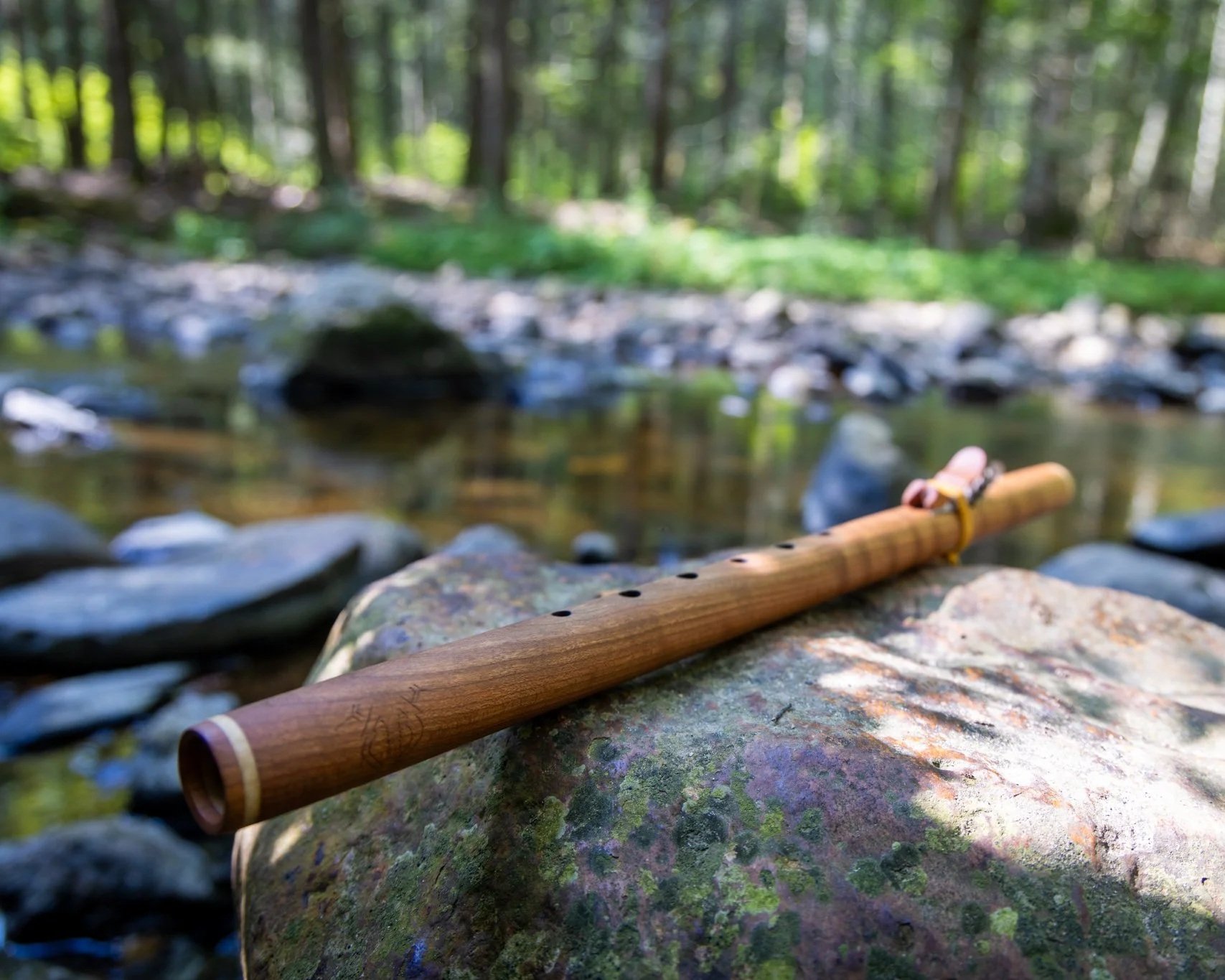A wooden flute rests on a mossy rock beside a shallow creek in a forest, with blurred trees and greenery in the background.
