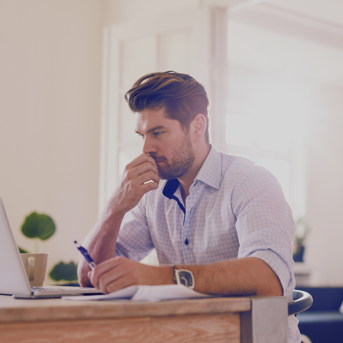 Man sitting at a desk, looking thoughtful and concentrating on his laptop, with sunlight streaming in from a window behind him.