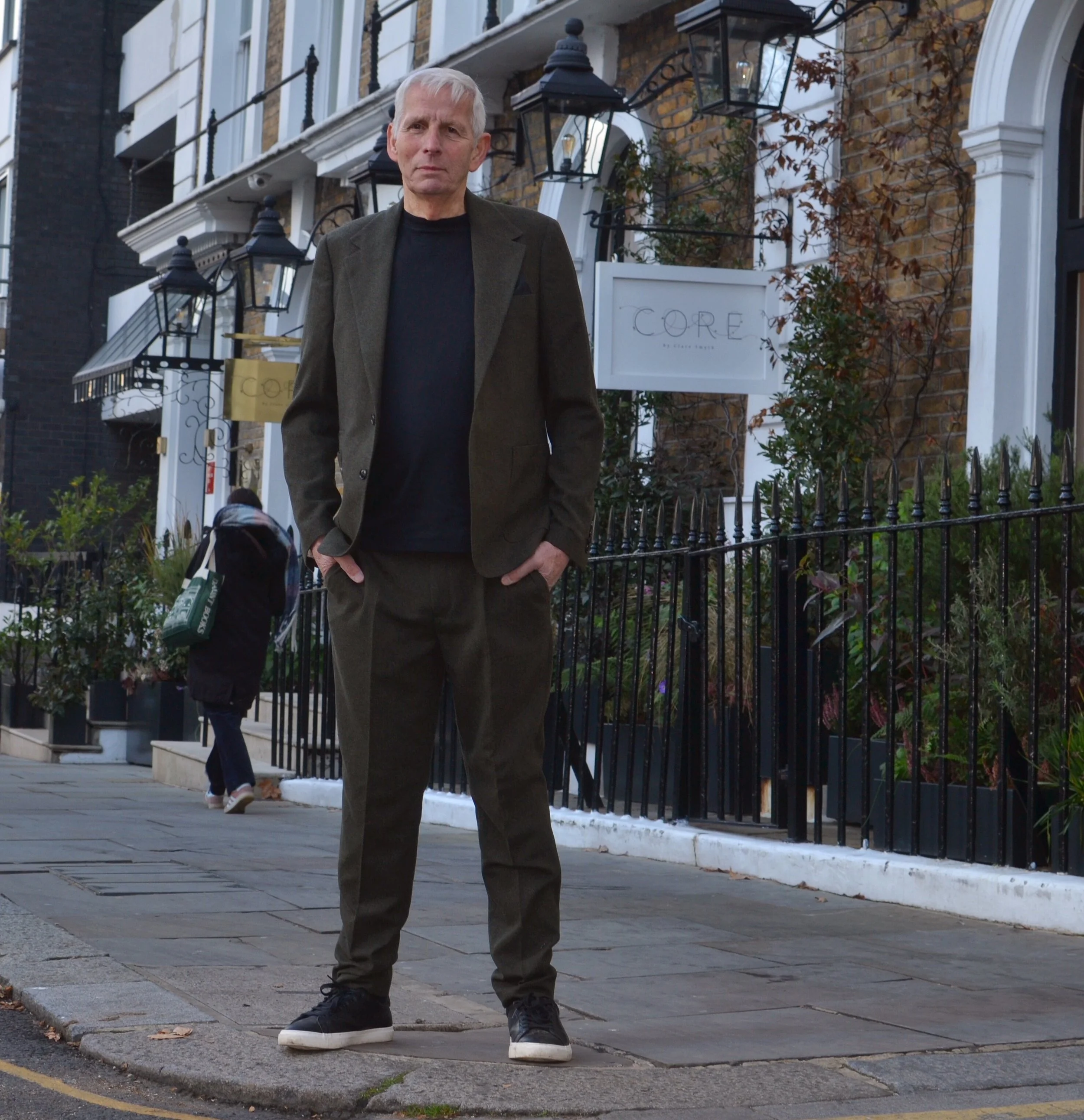 A man standing on a city sidewalk in front of a row of townhouses with black fences and lantern-style streetlights. He is wearing a dark green blazer, black shirt, and dark pants, looking directly at the camera with hands in his pockets.