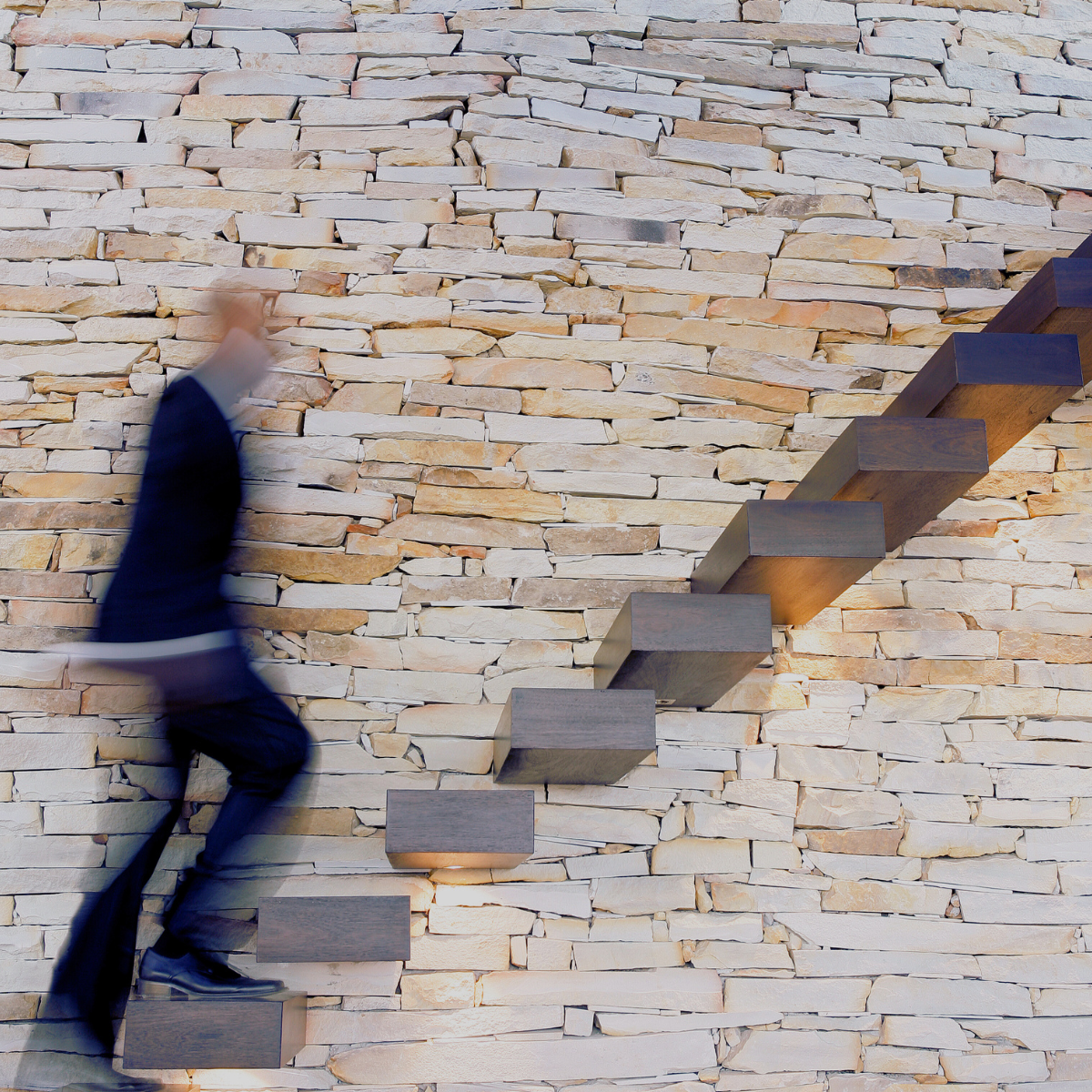 Person in suit climbing a modern staircase against a stone brick wall.