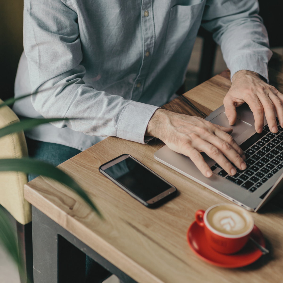 Person in a light gray shirt working on a silver laptop at a wooden table in a cafe, with a smartphone and a cup of coffee nearby.