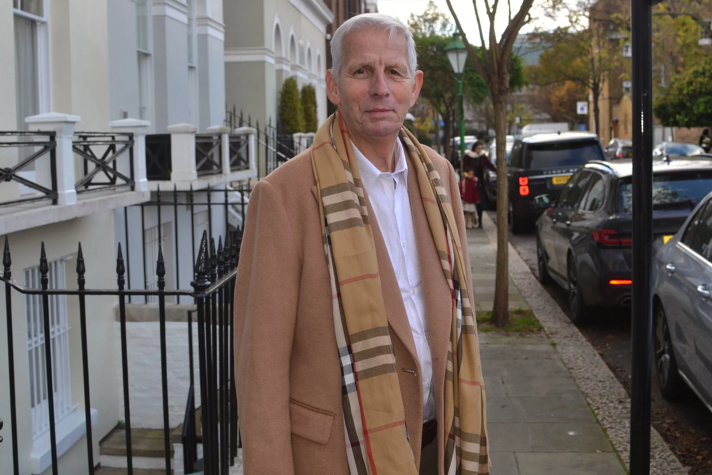 An older man with white hair standing on a city sidewalk, wearing a beige coat and a beige plaid scarf, with parked cars and a row of trees in the background.
