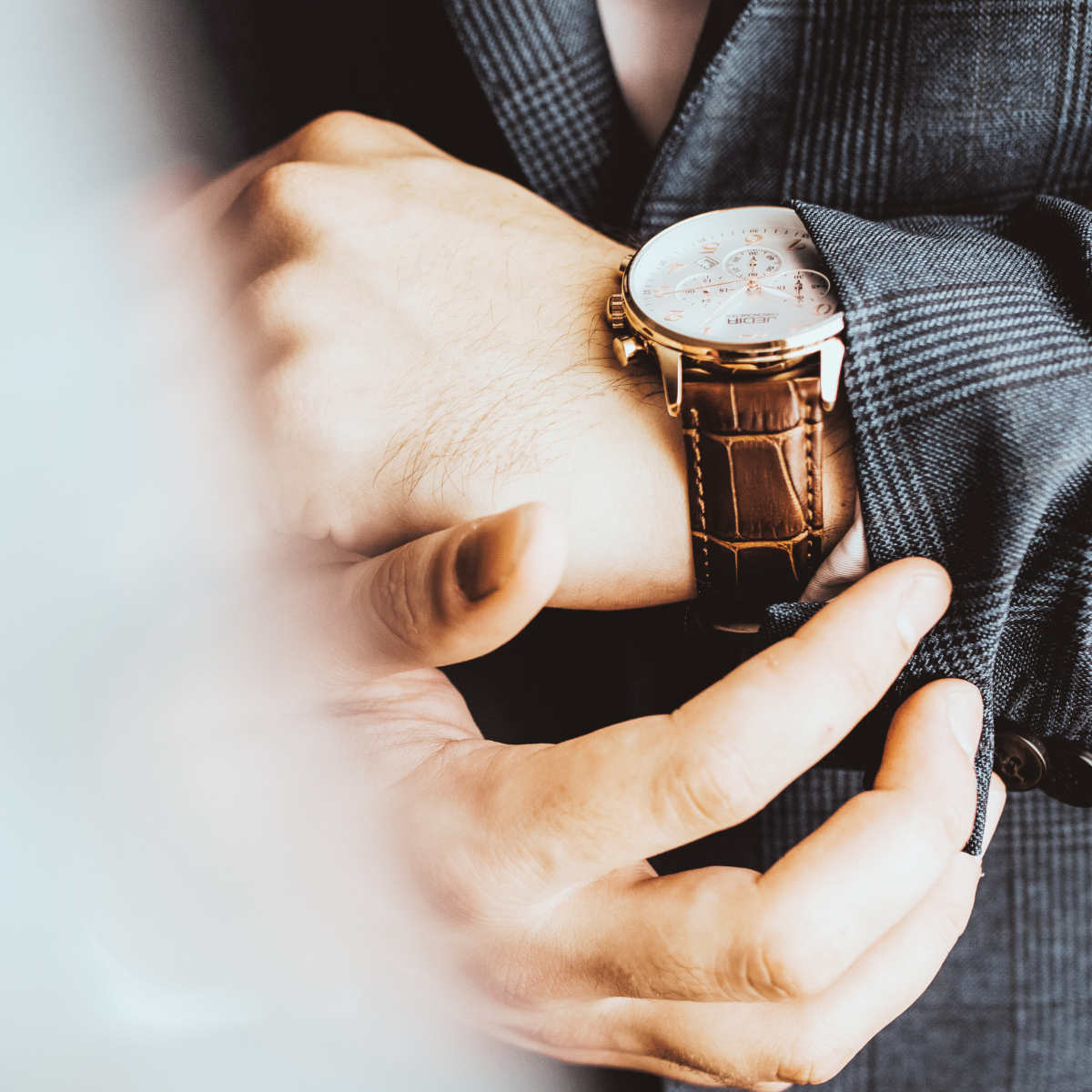A man wearing a checkered blazer and a large watch with a brown leather strap adjusts his cuff.