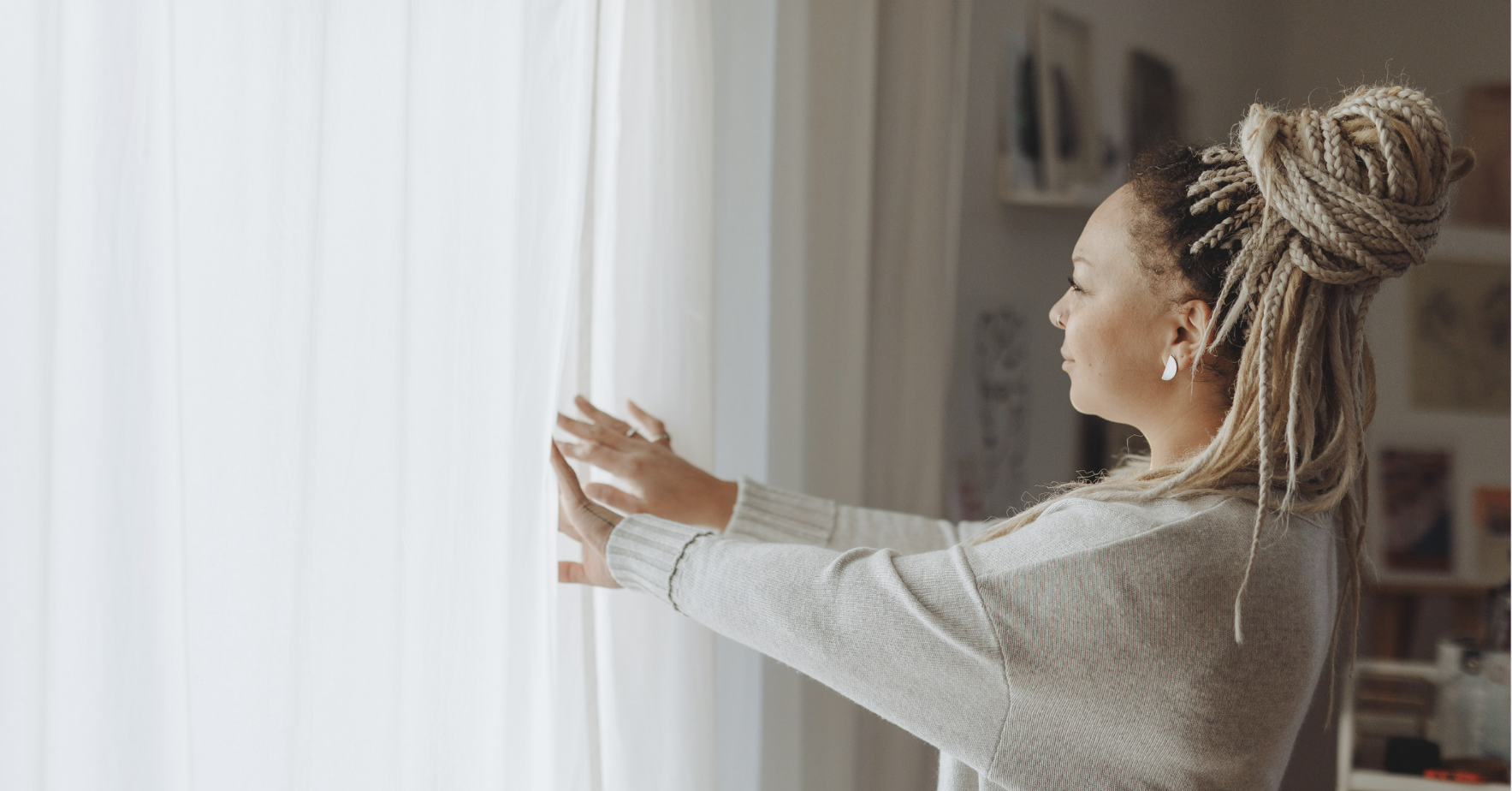 Woman wearing a white sweater opening white curtains.