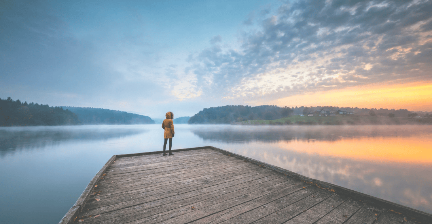Women standing at the end of a dock wearing a brown coat and looking out over a calm lake and a sunrise.
