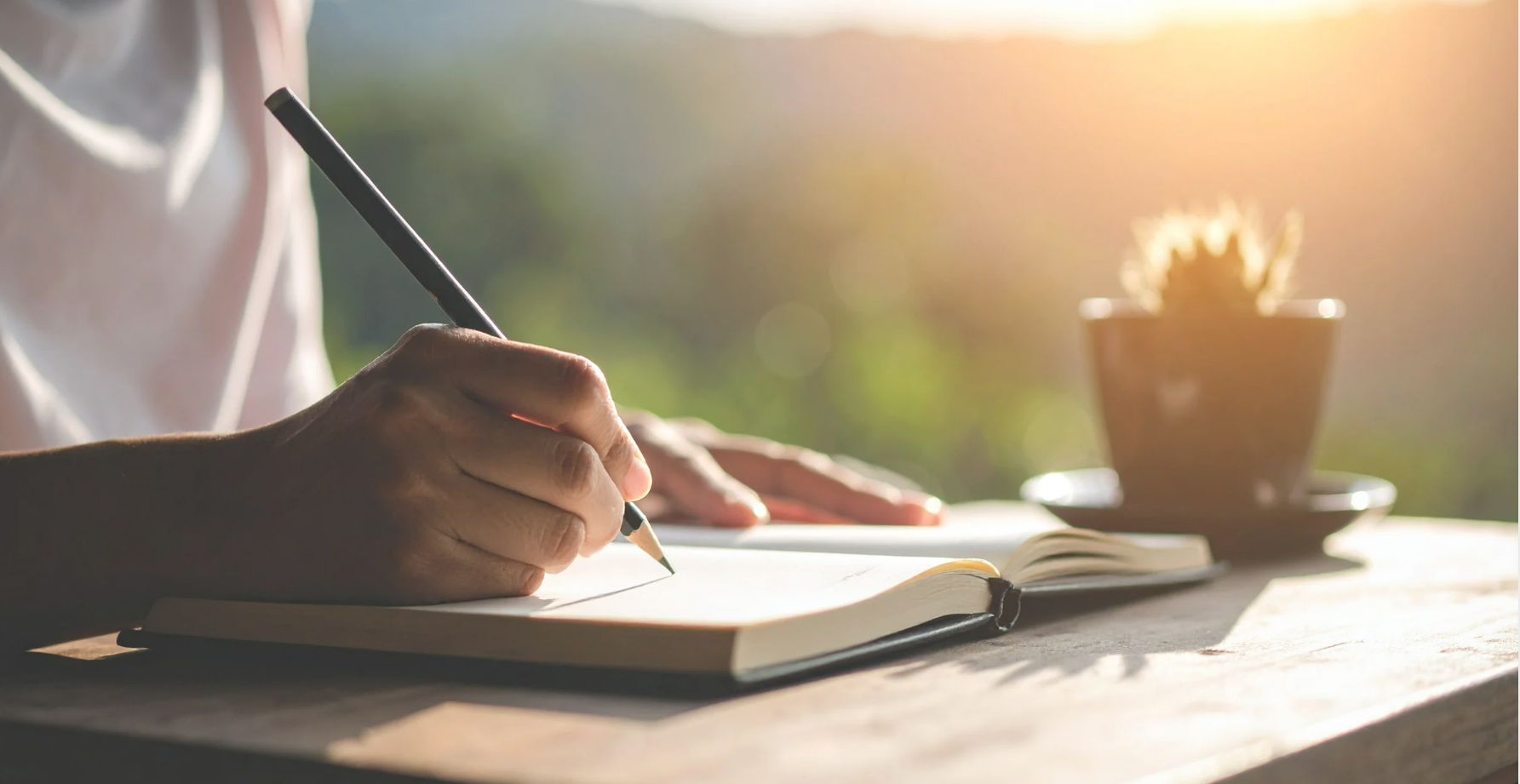 Person sitting at a desk and writing in a journal with a pencil.