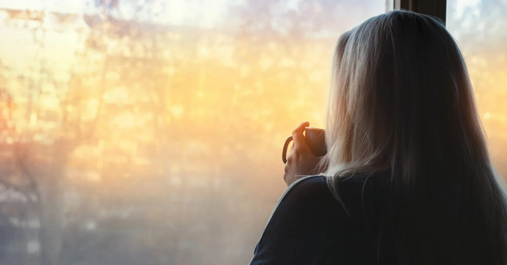 A woman holding a mug and looking out of a window in the early morning
