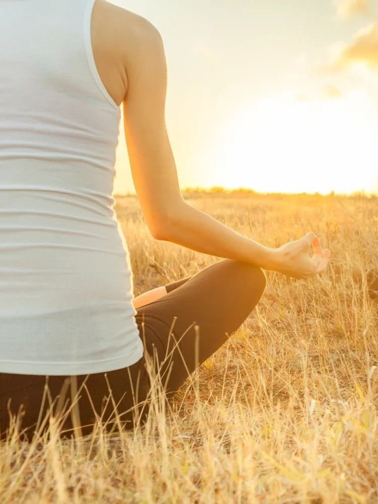 Person practicing meditation outdoors at sunset, sitting in a cross-legged position in a grassy field.