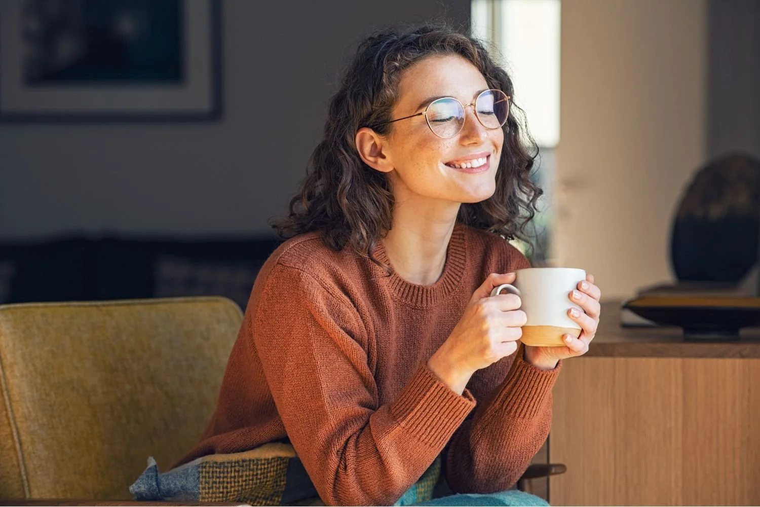 A woman with curly brown hair, wearing glasses and a brown sweater, smiling with eyes closed while holding a coffee mug inside a cozy room.