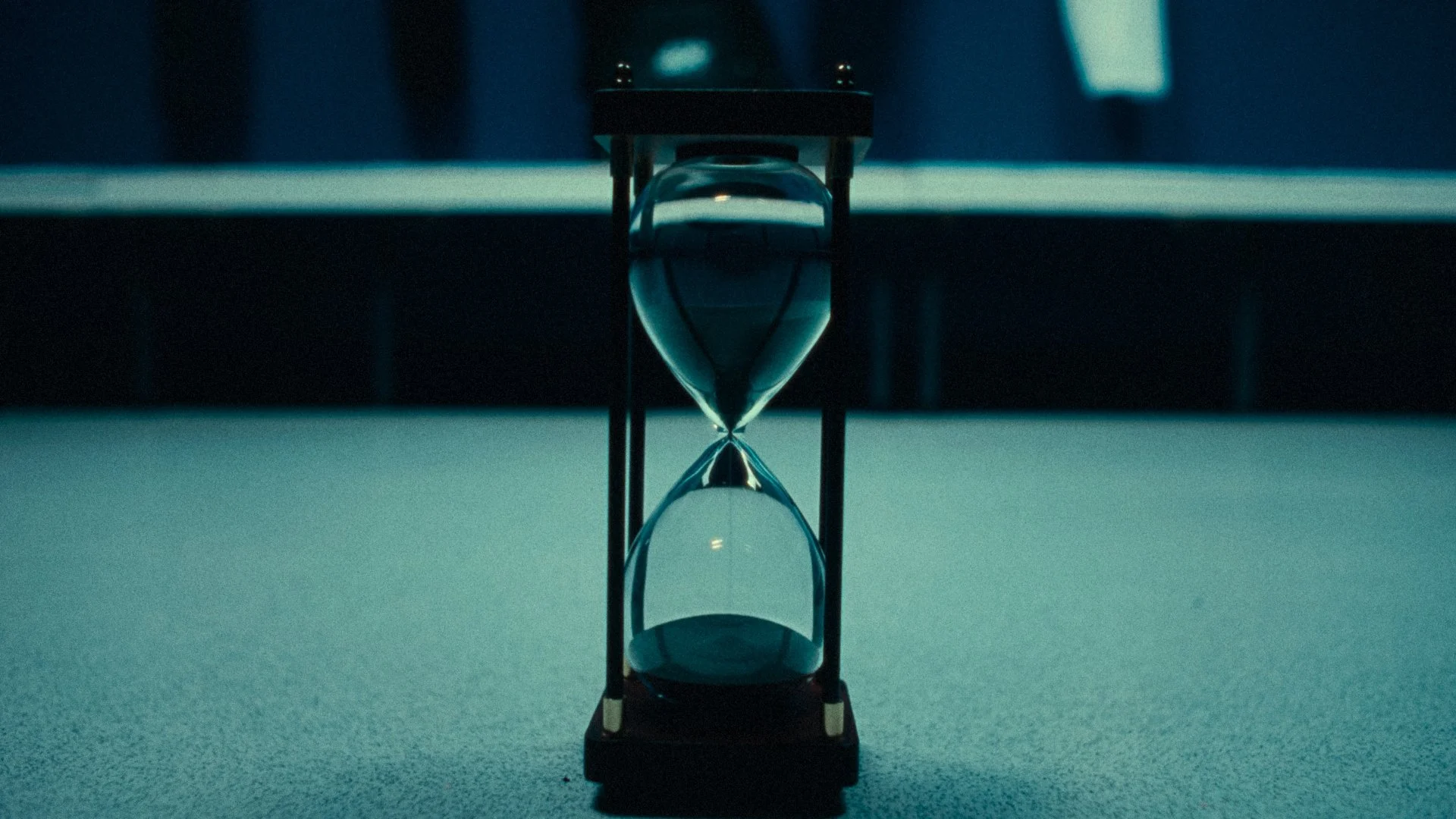 Close-up of a glass hourglass with blue sand, placed on a textured surface with a dark, blurred background.