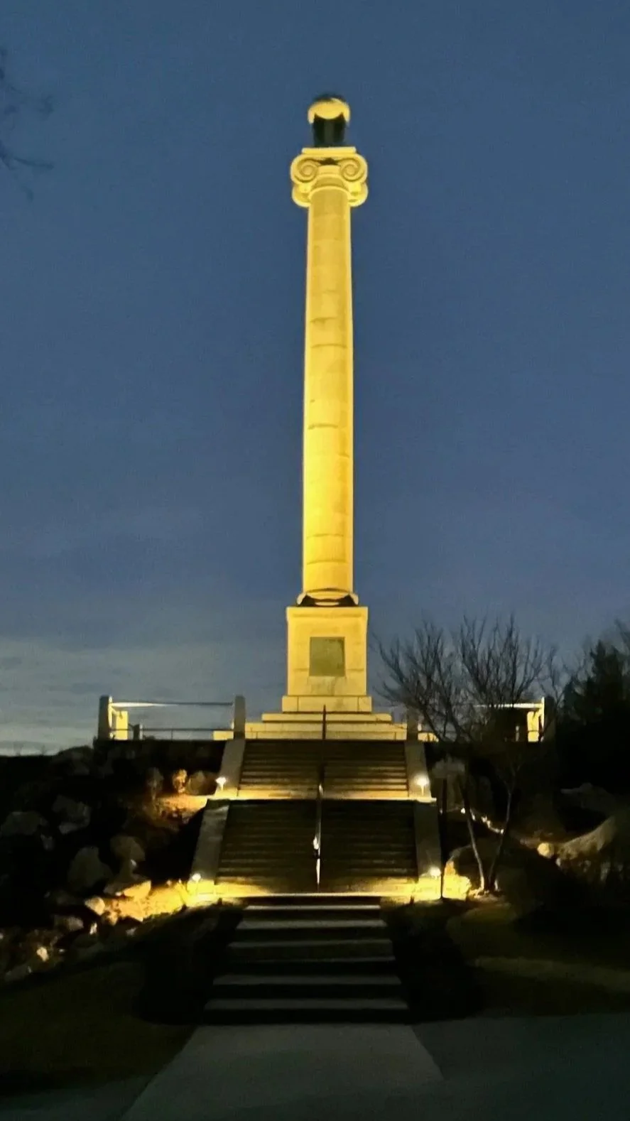 A tall monument illuminated at night, with stairs leading up to it, flanked by rocks and trees.