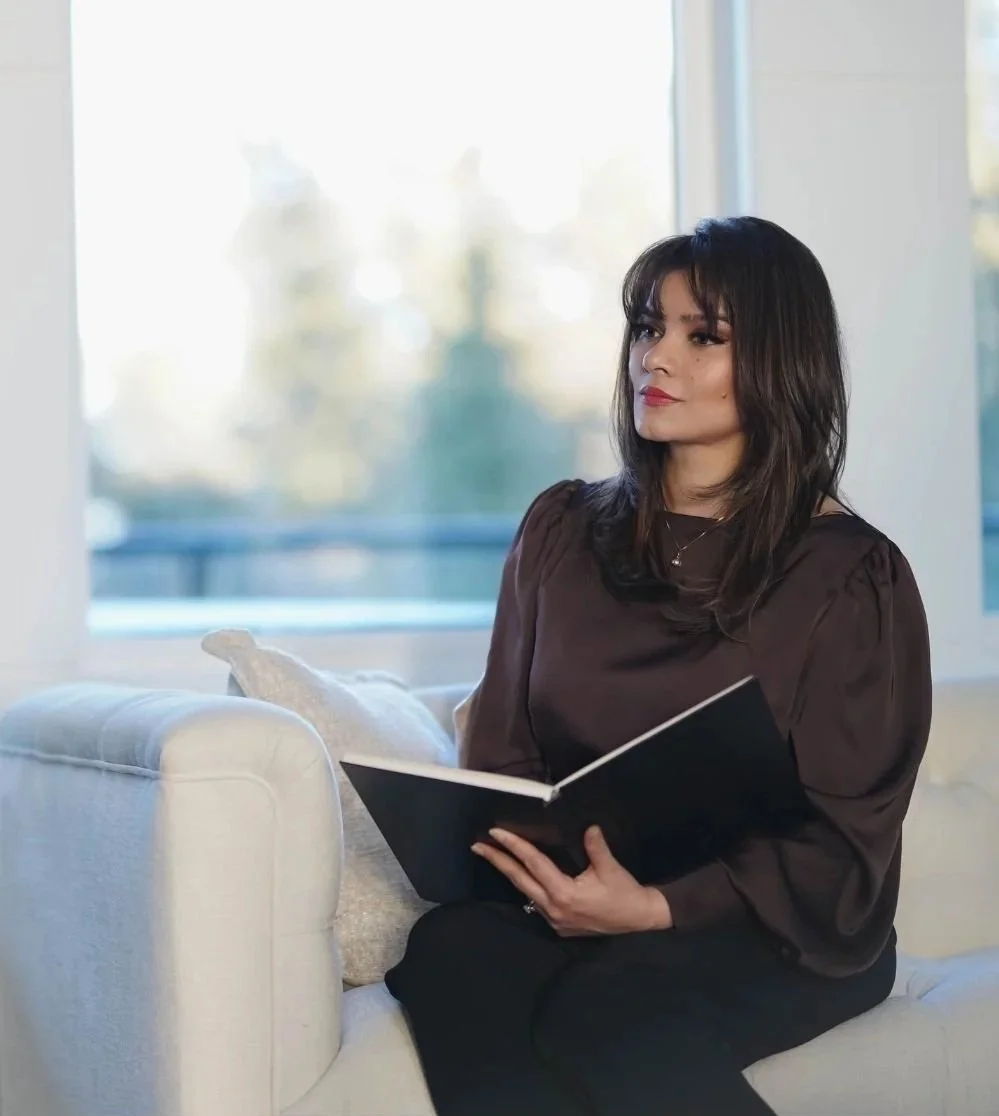 Ayesha sitting on a white couch holding a notebook at Art of Becoming Counselling