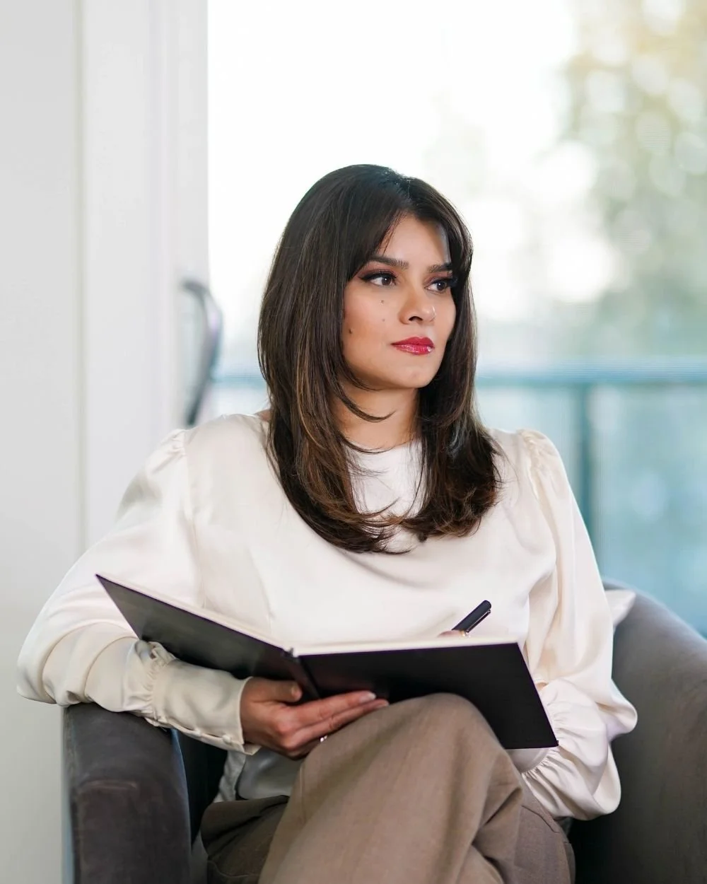 Ayesha sitting on a gray couch holding a notebook at Art of Becoming Counselling