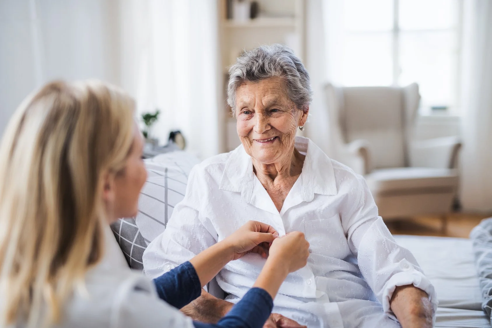An elderly woman sitting on a bed, smiling and engaging in conversation with a caregiver who is gently holding her hand in a bright, cozy room.