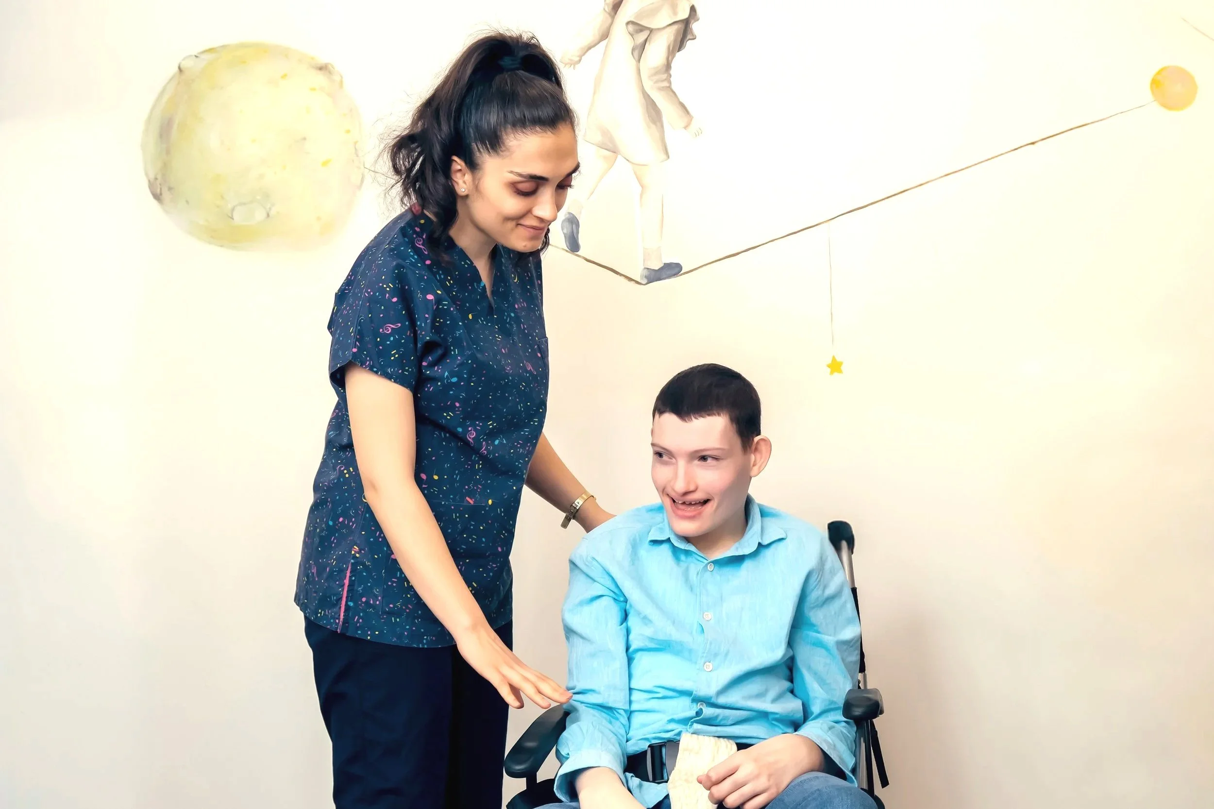 A young woman with dark hair in a ponytail is standing next to a seated young man in a wheelchair. They are in a bright room with a space-themed mural on the wall, including a large moon, a spacewalker, stars, and planets. The woman is smiling gently while the young man, who has short dark hair, is smiling with his eyes slightly closed. The young man is holding a sandwich in his right hand.