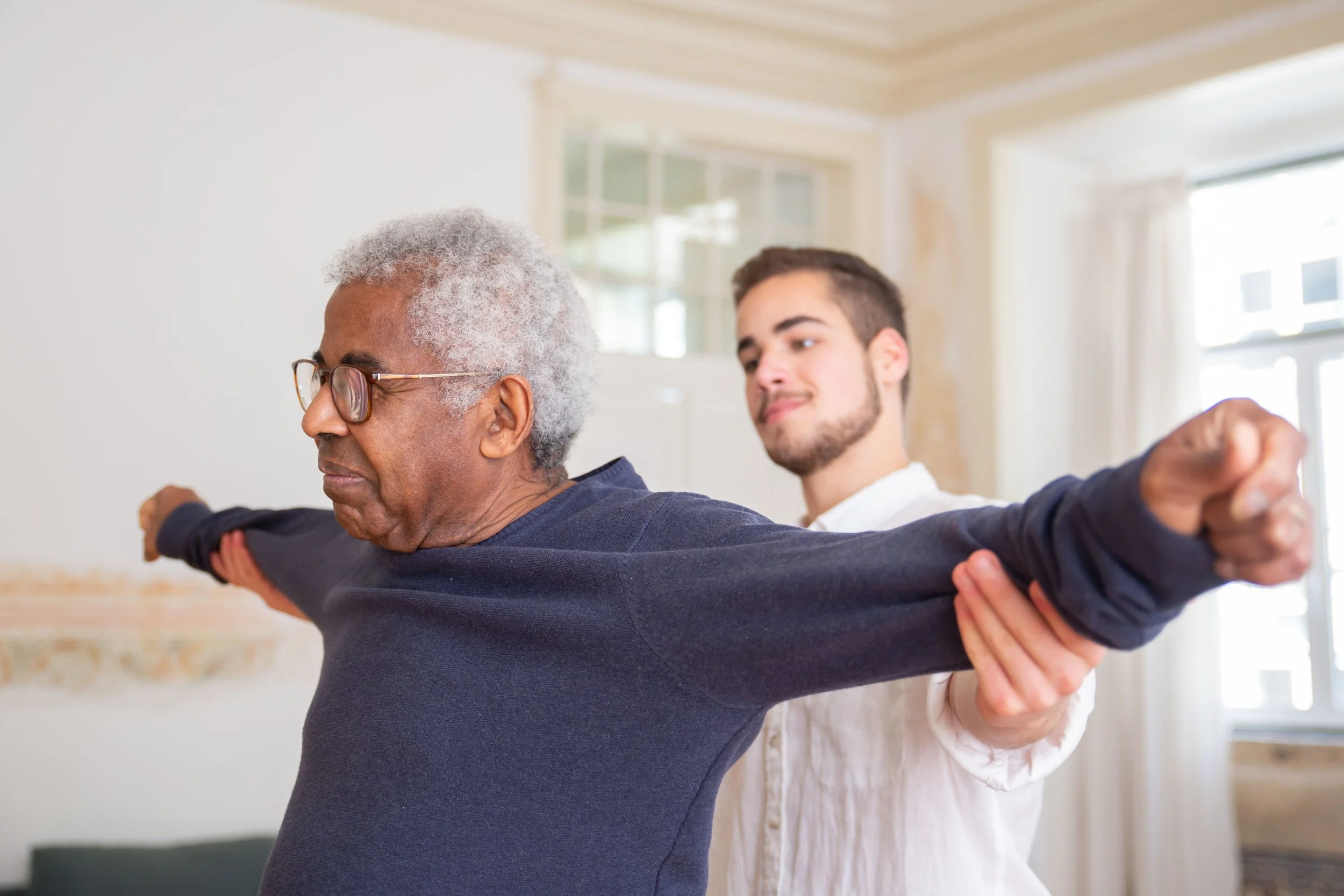 An elderly man and a caregiver practicing therapeutic exercises together indoors, with arms extended.