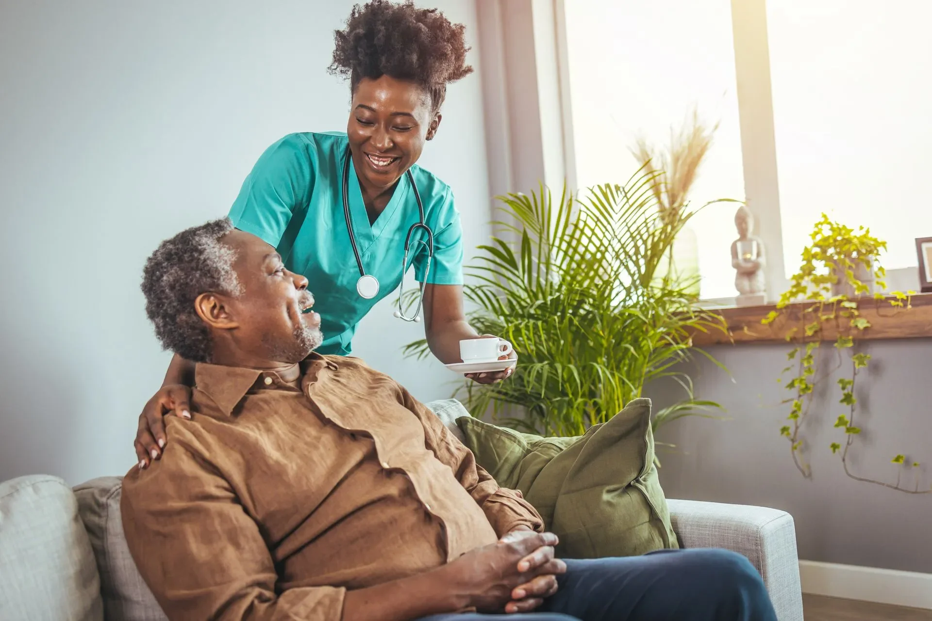A nurse/caregiver feeding an elderly man at home sitting on a couch in a well-lit room with plants and decorative items.