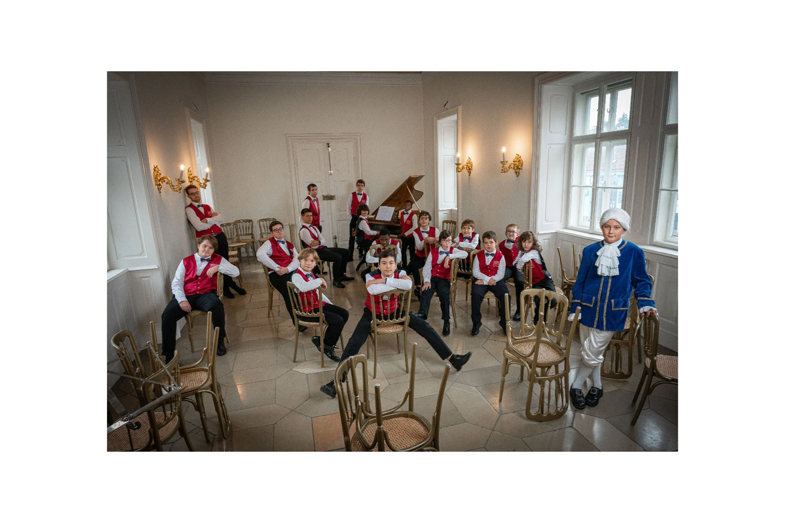 A group of young boys in formal attire, sitting on chairs in a spacious room with large windows. One boy stands dressed in historical costume, and there is a woman with a white bonnet and blue coat standing near the chair in the foreground. The room 