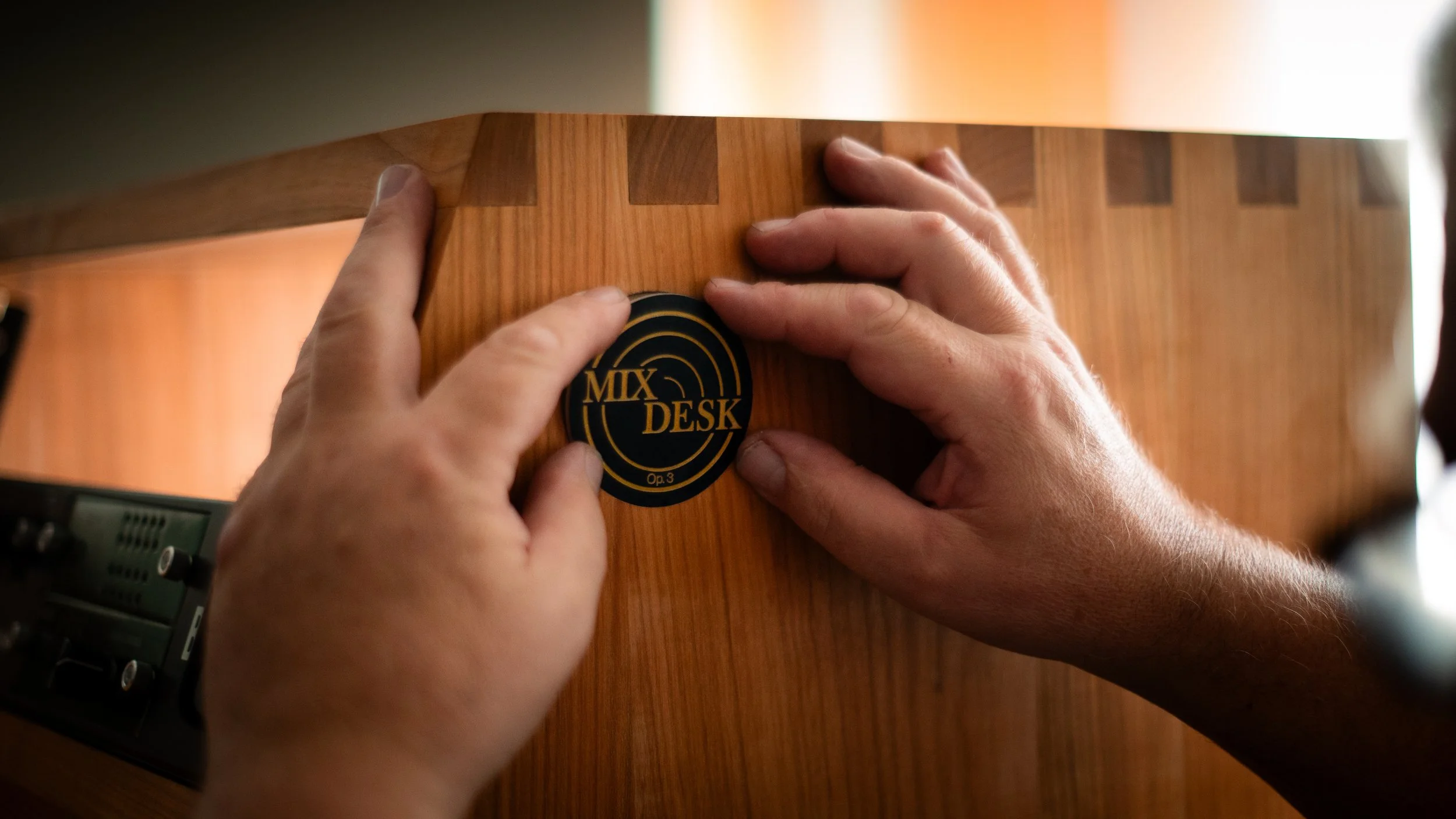 Hands placing a black and gold sticker that says "MIX DESK" on a wooden music mixing console.