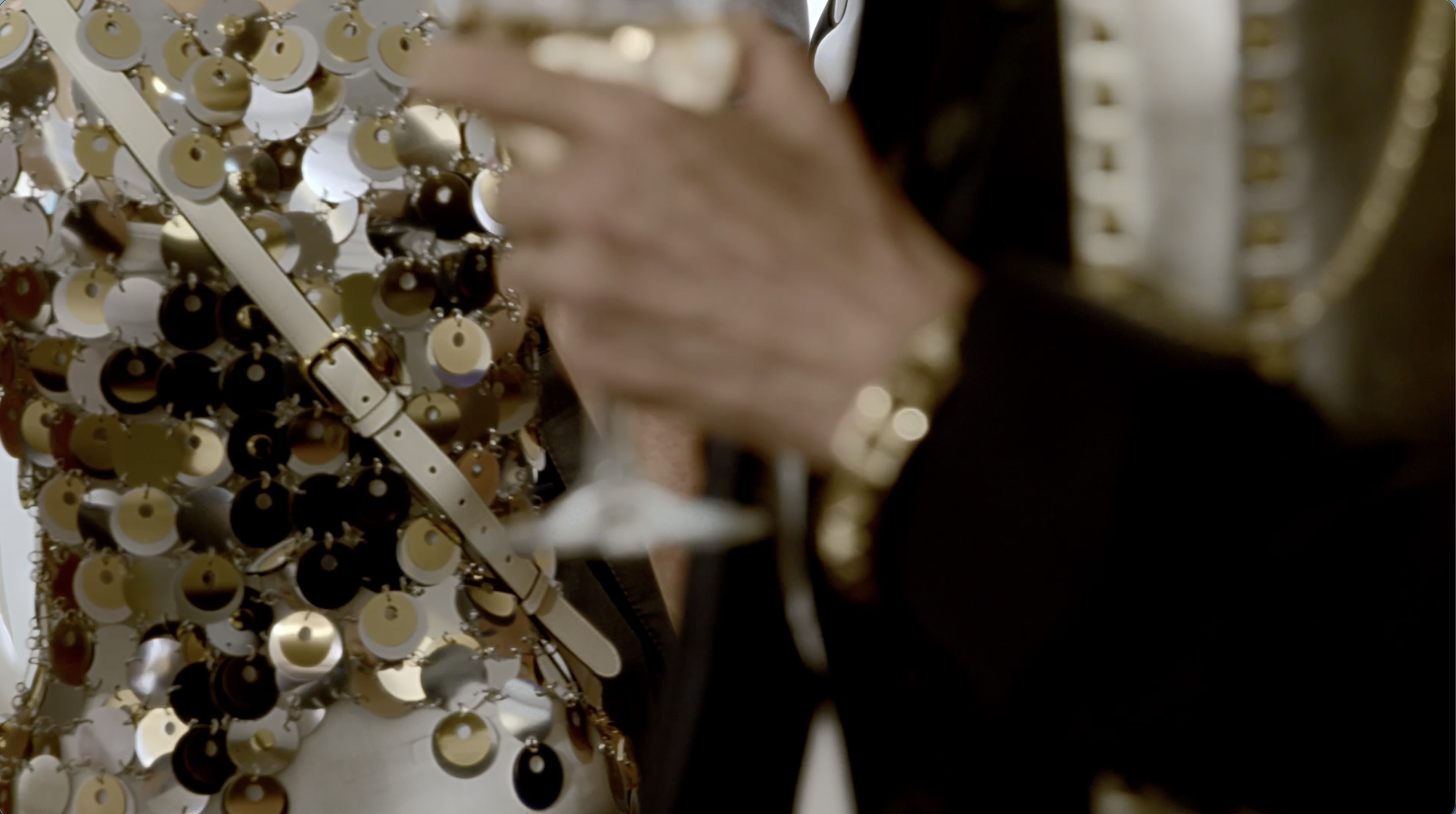 Glamorous close-up of a man in a dark suit holding champagne and wearing gold jewellery, with a woman in an oversized gold sequin dress shimmering in the background.